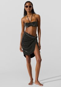 Fekete csillogó bikinifelső csomós részlettel, hozzá passzoló, oldalhasítékos, ráncolt szoknyával. Kiegészítők: nagyméretű napszemüvegek.