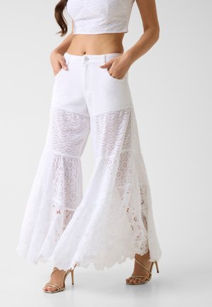 Femme portant un pantalon blanc à jambes larges avec tissus superposés en dentelle et en œillets, associé à des escarpins dorés à brides, debout les mains dans les poches.