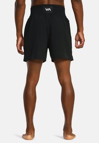 Mannelijke model staat blootsvoets met zijn rug naar de camera, gekleed in zwarte sportshorts met een VA-logo op de tailleband.