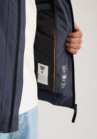 Hand holding open navy jacket showing black padded interior with label and icons indicating windproof, water repellent, and padded features.