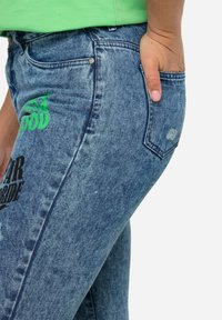 Vaqueros de mezclilla azul con una textura suave, bolsillos laterales y texto impreso en negro y verde. Zonas desgastadas y detalles de costura visibles.