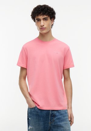 Ung mand med krøllet hår iført en lyserød T-shirt med rund hals og blå slidte jeans, stående med den ene hånd i lommen.