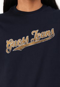 T-shirt in cotone blu navy con una grande grafica testurizzata in oro e bianco "Guess Jeans" sul petto e collo rotondo.