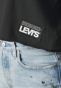 Étiquette de chemise noire Levi's sur une personne portant un jean en denim bleu clair avec des rivets métalliques près de la poche avant.
