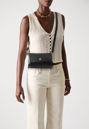 Borsa a tracolla nera in pelle con design a ribalta, accenti dorati e tracolla removibile, indossata con un top lavorato a maglia beige testurizzato e pantaloni chiari.