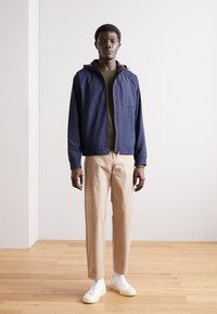 Jachetă cu glugă de culoare bleumarin, cu accente roșii, cămașă verde, pantaloni bej și adidași albi. Design minimalist, textură fină și siluetă ajustată.