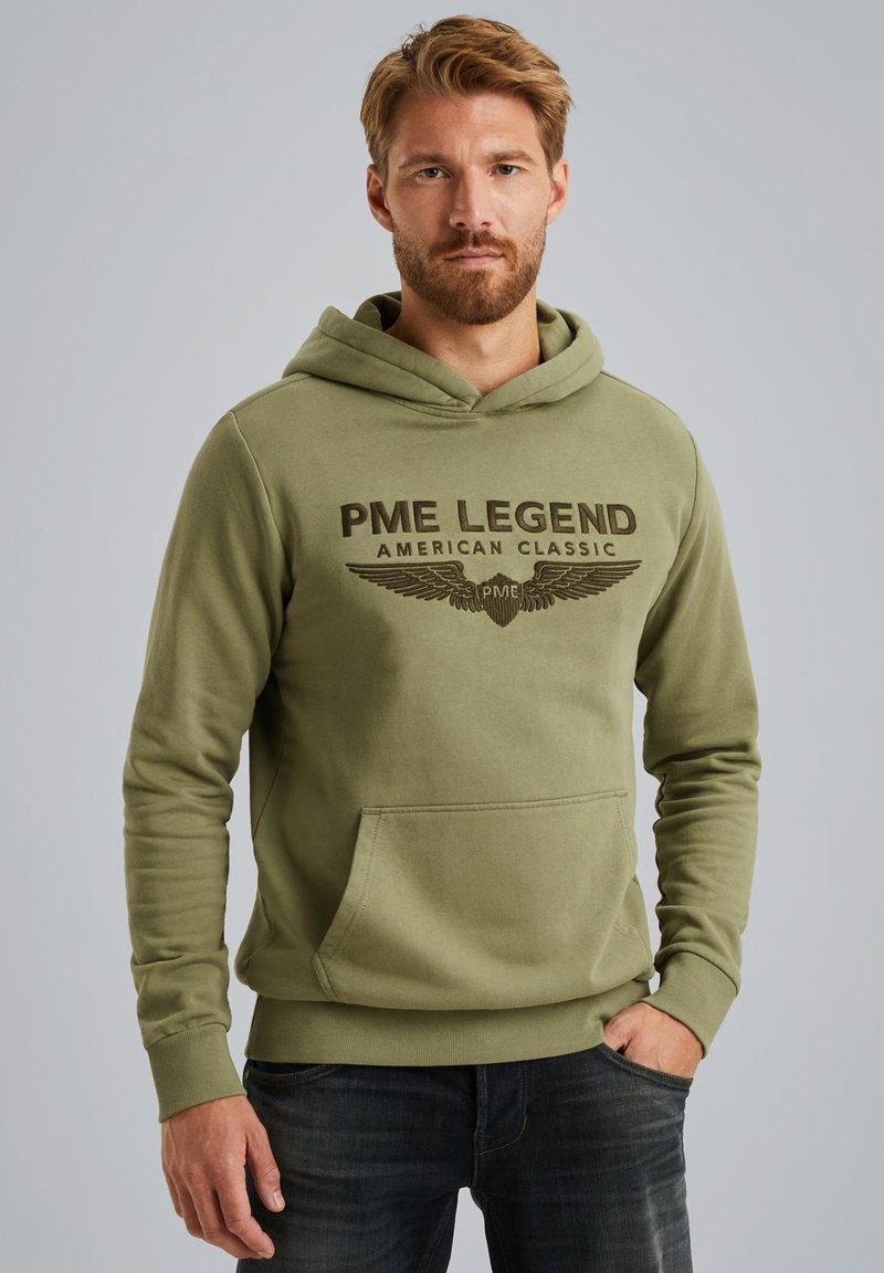 Olive green hoodie made of soft fabric featuring a front pocket and a logo print with winged accents. Model wears the hood up.