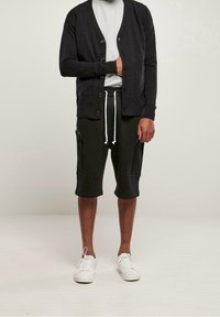 Svart kofta med knappar, bärs över en vit t-shirt. Kombinerad med mörka shorts som har dragkedjefickor och vita sneakers.