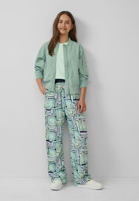 Muntgroene bomberjack met geribbelde boorden, gecombineerd met geprinte broeken met vierkanten in pastelkleuren. Witte sneakers maken de look af.