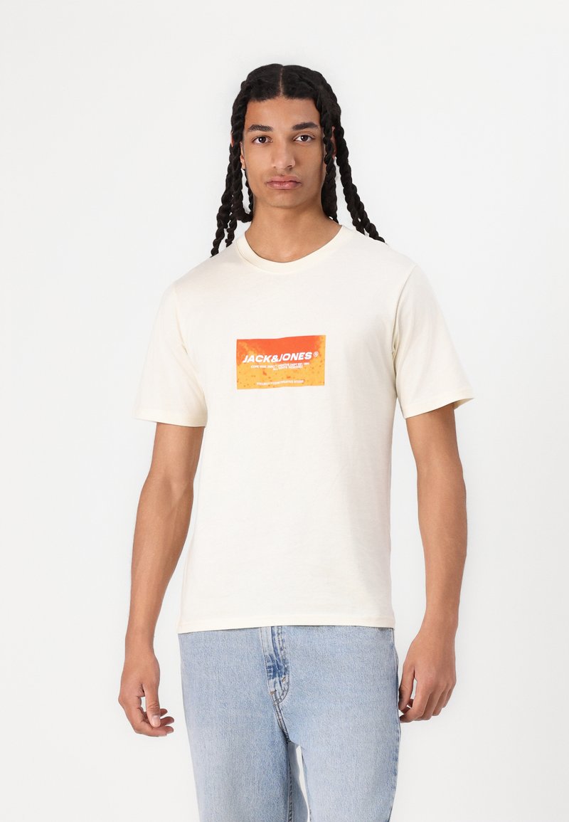 Crème katoenen t-shirt met een rechthoekige oranje logopatch met witte tekst, korte mouwen en een klassieke ronde hals.