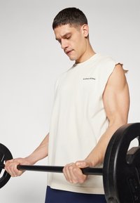 Cream sleeveless workout top with raw edges and 'Björk Borg' logo, held while lifting a black weight bar. Plain background.