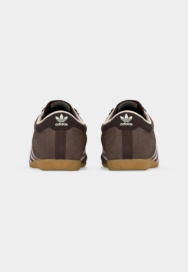 Par de zapatillas Adidas marrones con suelas beige, vistas desde atrás, mostrando el logo de Adidas y tres rayas en rosa pálido en los costados.