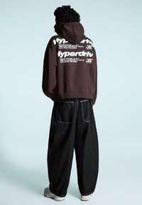 Brązowa bluza z kapturem z białym napisem na plecach, zestawiona z luźnymi czarnymi dżinsami. Do stroju dołączone są białe trampki. Tło jest jasnoniebieskie.