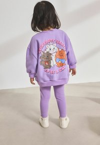 Lila pulóver, hátán mesefigurás macskákkal, lila leggings-szel párosítva és fehér bokacsizmával. Puha anyag, lazán illeszkedő fazon.