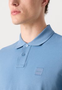 Gaiši zils polo krekls, kas izgatavots no teksturēta auduma. Tam ir klasiskas formas apkakle, divi pogas un kvadrātveida logo plāksne uz krūtīm.