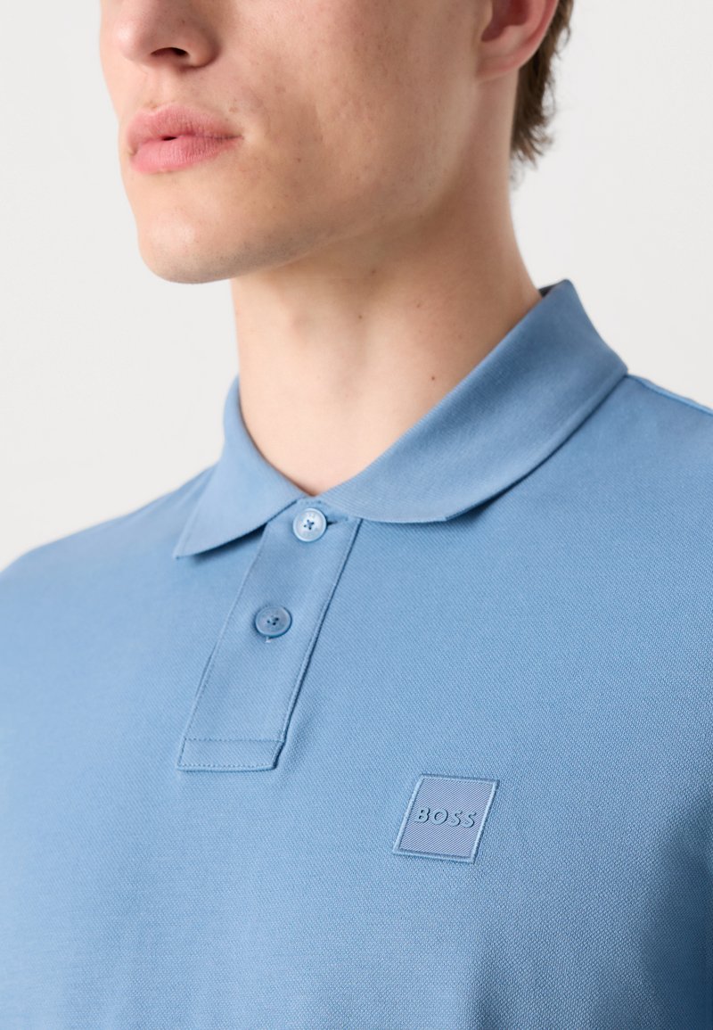 Polo de color azul claro hecho de tejido texturizado. Presenta un collar clásico, dos botones y un parche cuadrado con el logo en el pecho.