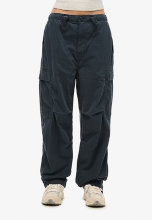 Personne portant un pantalon cargo noir ample avec des poignets élastiques et des baskets beiges, debout devant un fond blanc uni.