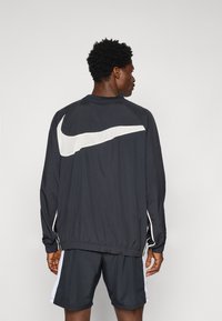 Svart Nike-jakke med hvit logo, med elastiske mansjetter og hem, parret med svarte shorts. Behagelig tekstur, atletisk passform.