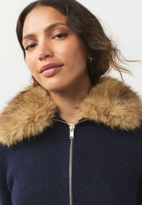 Giacca con zip blu navy con colletto in pelliccia ecologica beige. Cerniera metallica con tessuto strutturato. Vista ravvicinata che mette in risalto il dettaglio del colletto.