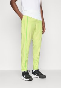 Pantalones de chándal verde claro con franjas laterales en neón verde contrastante, fabricados con una tela suave. Combinados con zapatillas deportivas negras con acentos en blanco.