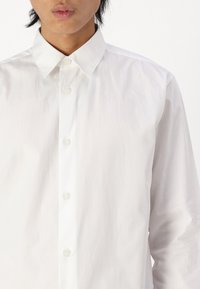 White button-up shirt with a spread collar, featuring a smooth texture and five buttons down the front. Simple, classic design.