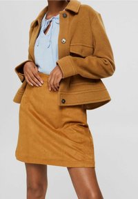Femme portant un chemisier bleu clair à col noué, une veste en laine couleur camel avec boutons, et une jupe assortie en daim camel.