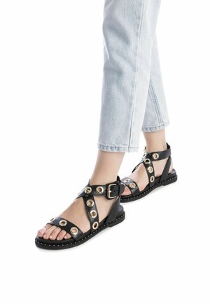 Sandali in pelle nera con accenti circolari dorati, cinturino con fibbia regolabile, suola piatta e bordo borchiato; indossati con jeans blu chiaro.