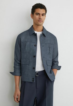 Mand med krøllet hår iført en grå knappet jakke over en hvid skjorte og mørke bukser, stående med den ene hånd i lommen mod en hvid væg.