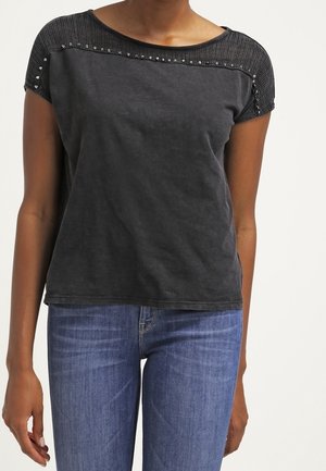 Kvinde iført en sort kortærmet top med teksturerede skuldre og nitte detaljer, kombineret med blå denim jeans.
