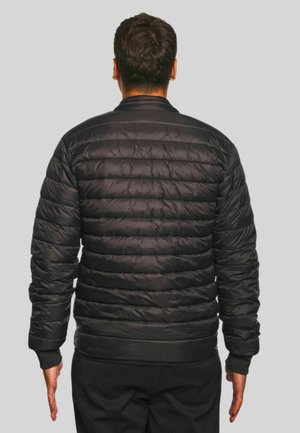 Hombre de espaldas con una chaqueta acolchada negra y pantalones negros, sobre un fondo gris liso.