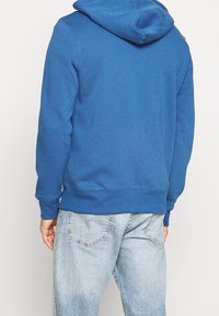 Personne portant un sweat à capuche bleu uni et un jean en denim bleu clair, vue de dos sur un fond clair uni.