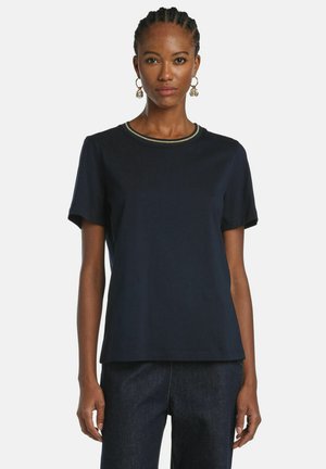 Frau mit geflochtenem Haar trägt goldene Creolen, ein schlichtes dunkelblaues kurzärmliges Hemd mit einem dezenten gestreiften Kragen und dunkle Jeans.