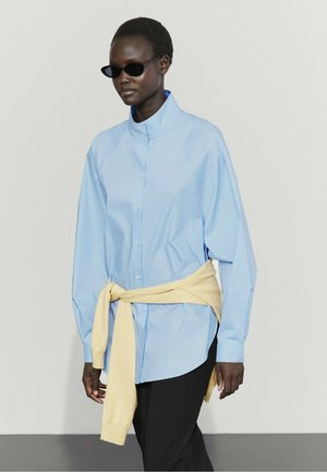 Femme portant une chemise bleu clair oversize boutonnée, un pull jaune noué autour de la taille, un pantalon noir et des lunettes de soleil noires sur fond uni.