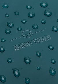 Teal vízálló anyag, melyen kiemelkedő „JOHNNY URBAN” logó található. Több vízcsepp látható, amely a textúrát és a sima felületet mutatja.
