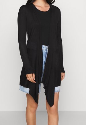 Femme portant un long cardigan noir ouvert devant avec des poches à l'avant, par-dessus un top noir et un short en jean bleu clair aux bords effilochés.