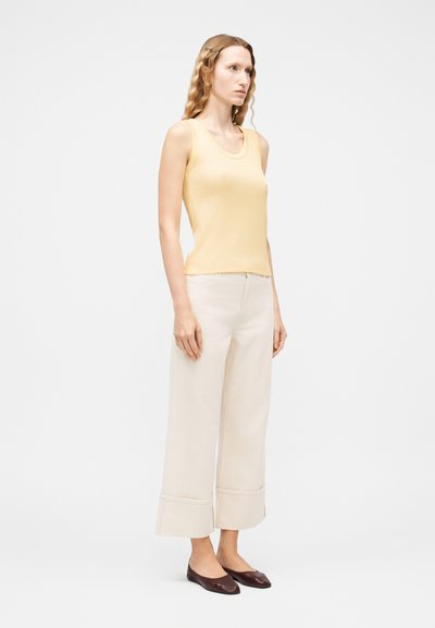 Γυναίκα με κυματιστά μαλλιά που φοράει ένα κίτρινο αμάνικο τοπ, παντελόνι σε κρεμ χρώμα με φαρδιές μπατζάκια και μανσέτες, και σκούρα καφέ μυτερά παπούτσια, στέκεται μπροστά σε λευκό φόντο.