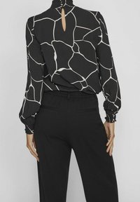 Černá blůza s bílým abstraktním vzorem, vysoký límec, otvor na zádech a nařasené manžety; kombinováno s černými kalhotami se bočními kapsami.