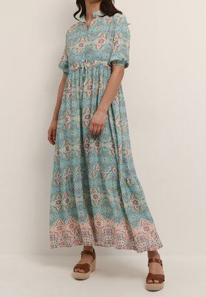 Femme portant une longue robe bleue à motifs, manches courtes bouffantes et sandales compensées marron sur un fond clair uni.