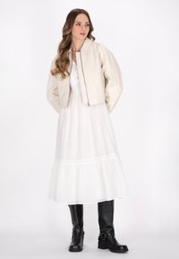 Bevergete leren jack over een witte, gelaagde maxidress met kantafwerking, gecombineerd met hoge zwarte laarzen met gespen.