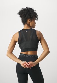 Črn športni crop top s prozornim mrežastim dizajnom, nastavljivimi naramnicami in elastičnim trakom z blagovno znamko na dnu; v kombinaciji s črnimi legicami.