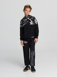 Czarna bluza z kapturem z białym wzorem marmurowym na g órze i kieszenią kangurką, w zestawie z czarnymi spodniami dresowymi z białym napisem i akcentami. Czarne trampki.