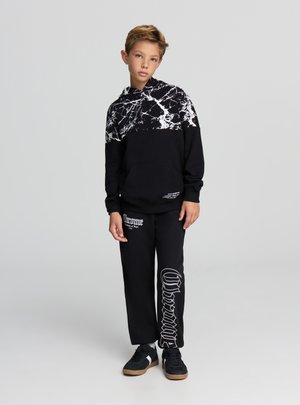Czarna bluza z kapturem z białym wzorem marmurowym na górze i kieszenią kangurką, w zestawie z czarnymi spodniami dresowymi z białym napisem i akcentami. Czarne trampki.