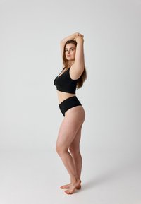 Vrouw staat op blote voeten, draagt zwart ondergoed, heeft haar armen omhoog en in elkaar gevlochten boven haar hoofd, kijkt vooruit tegen een effen achtergrond.