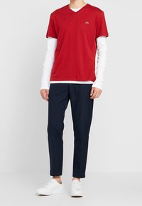 Maglietta rossa a V sovrapposta a una camicia bianca a maniche lunghe, abbinata a pantaloni blu navy e scarpe da ginnastica bianche. Design semplice, tessuto di cotone.