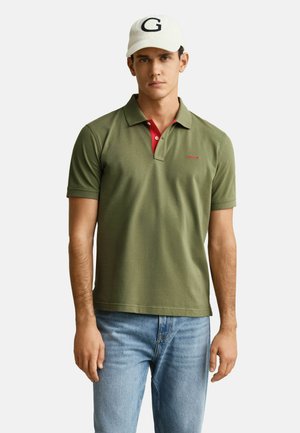 Jeune homme portant un polo vert olive avec des boutons rouges, un jean bleu clair et une casquette blanche avec une lettre G noire.