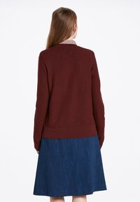Maglione lavorato a maglia bordeaux con scollatura tonda, polsini e orlo ribattuti, abbinato a una gonna di jeans lunghezza ginocchio. Dettaglio del colletto con motivo a quadri visibile.
