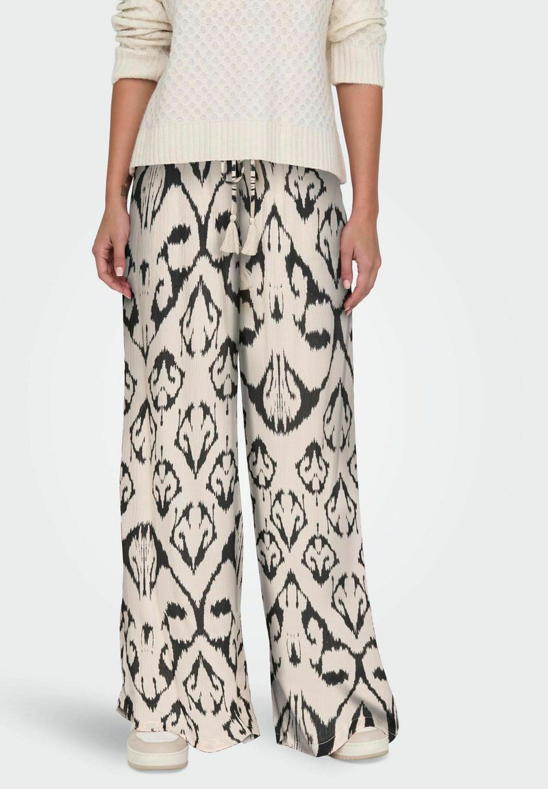 Femme portant un pantalon à jambes larges avec un motif abstrait noir et blanc, associé à un pull en maille crème et des baskets blanches.