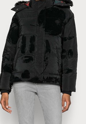 Schwarze gesteppten Steppjacke mit dezentem samtigem abstraktem Muster, Fronttaschen und Kapuze mit mehrfarbiger kariertem Futter, getragen mit hellgrauen Jeans.