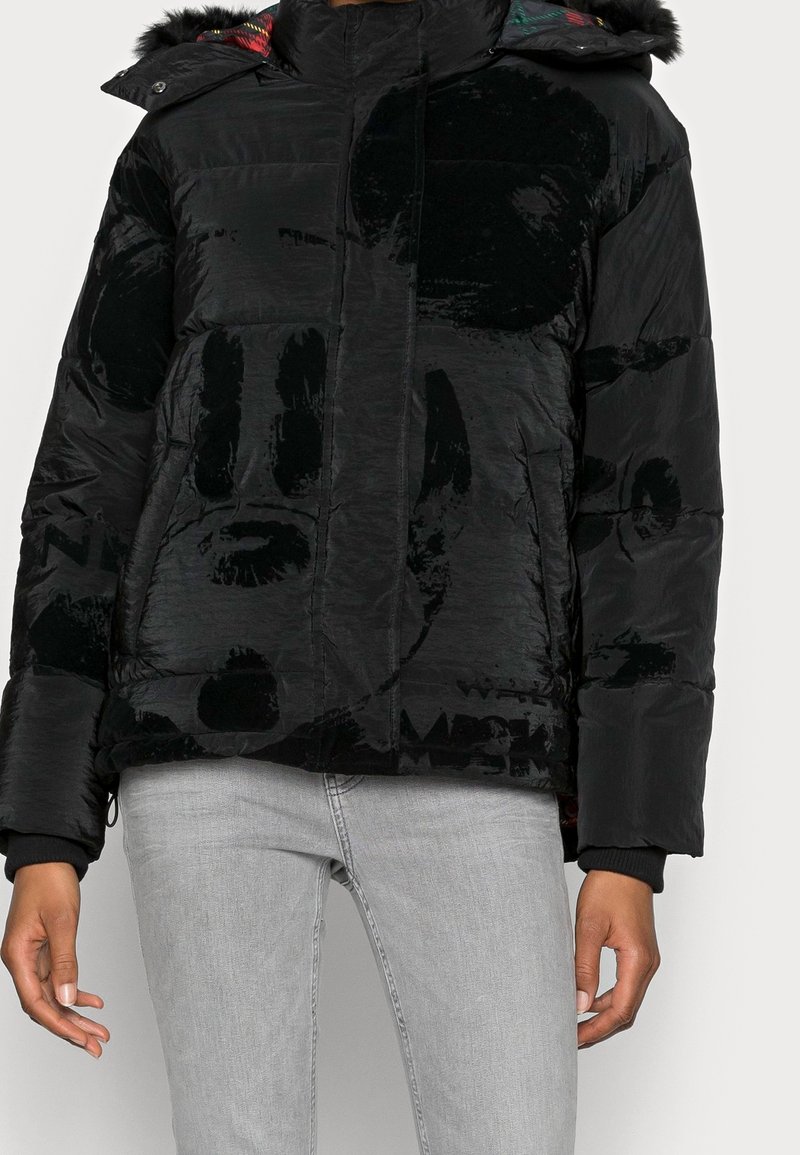 Veste matelassée noire avec un motif abstrait en velours subtil, poches avant, et une capuche doublée d'un tissu à carreaux multicolores, portée avec un jean gris clair.