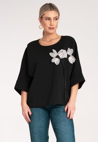 Czarna, oversize'owa bluzka z krótkim rękawem, z kwiatowym wzorem w bieli i czerni z przodu. Połączona z niebieskimi dżinsami.